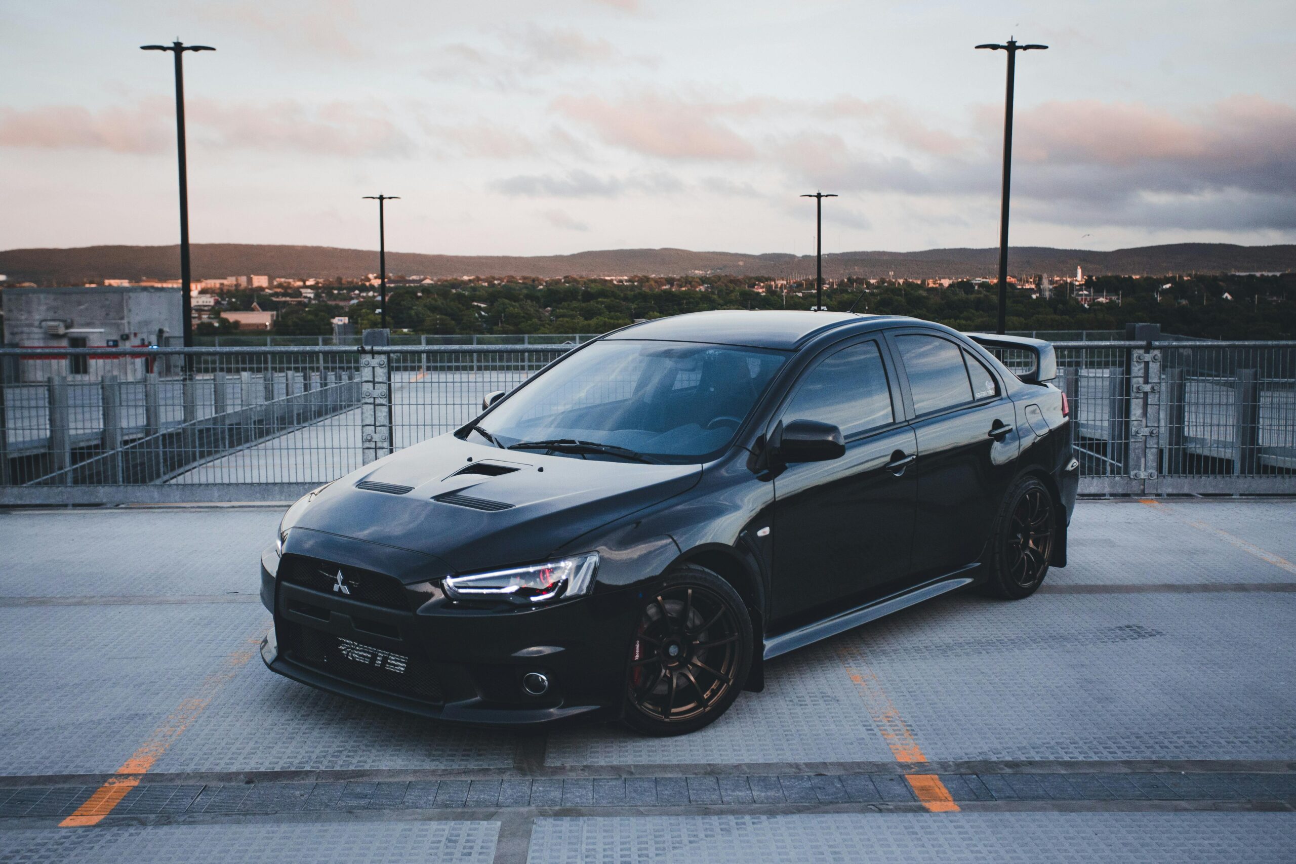 A stylish black car parked on a rooftop parking with a scenic sunset backdrop.