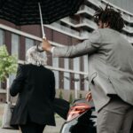 Two business professionals with an umbrella near a luxury car in an urban setting.