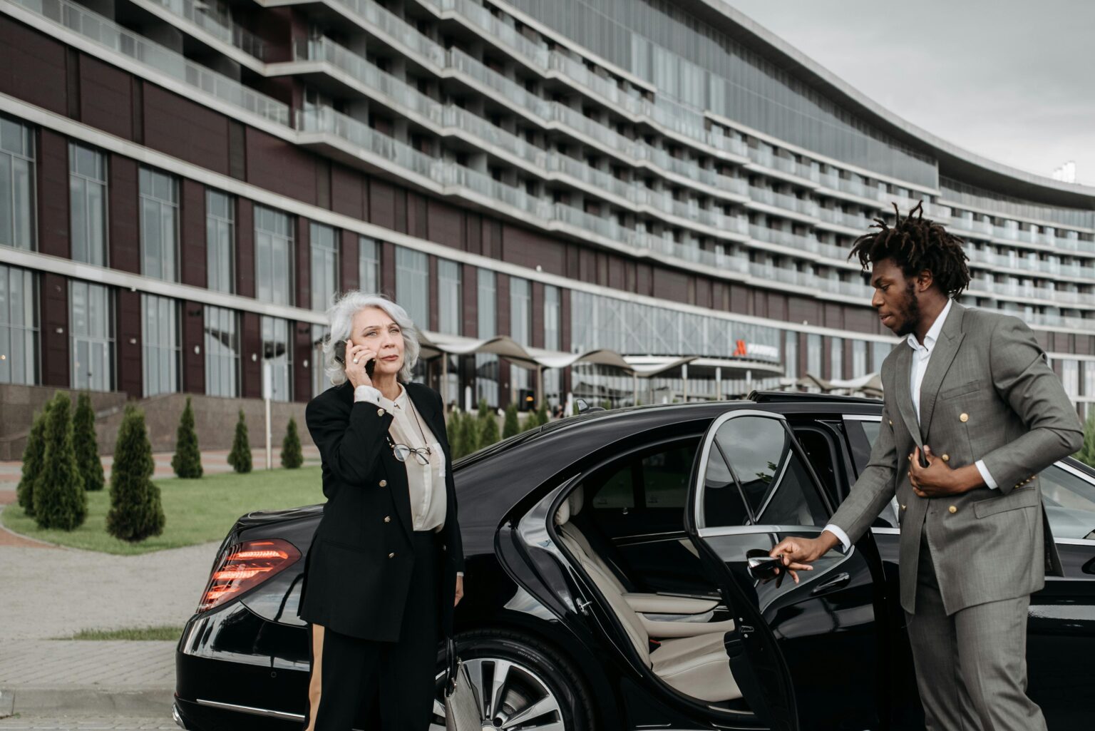 Senior female executive on phone with chauffeur holding car door open, modern architecture backdrop.